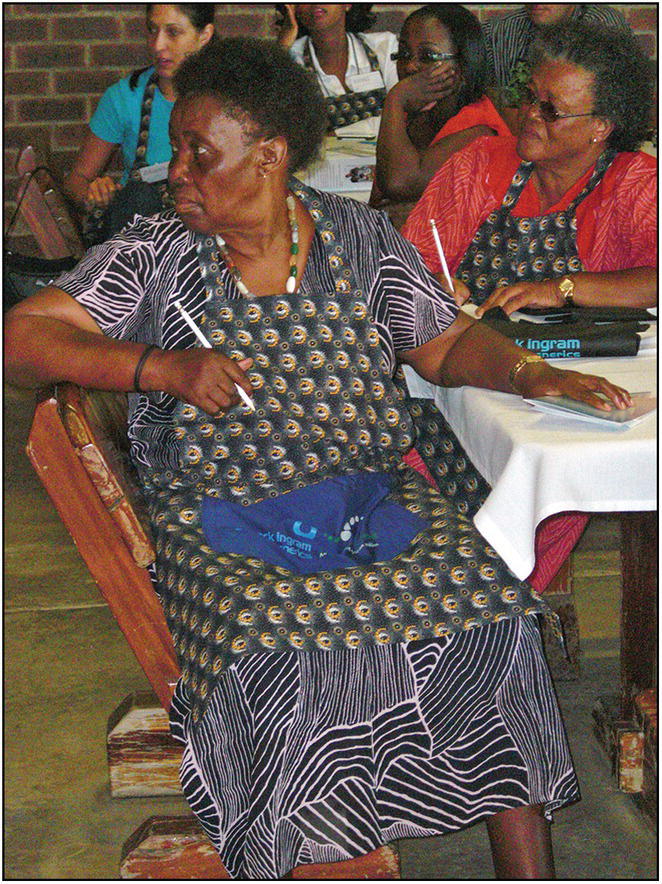 Photo of African women seated around a table and looking at a speaker (not in the image). Two of them are holding pencils and taking down notes.