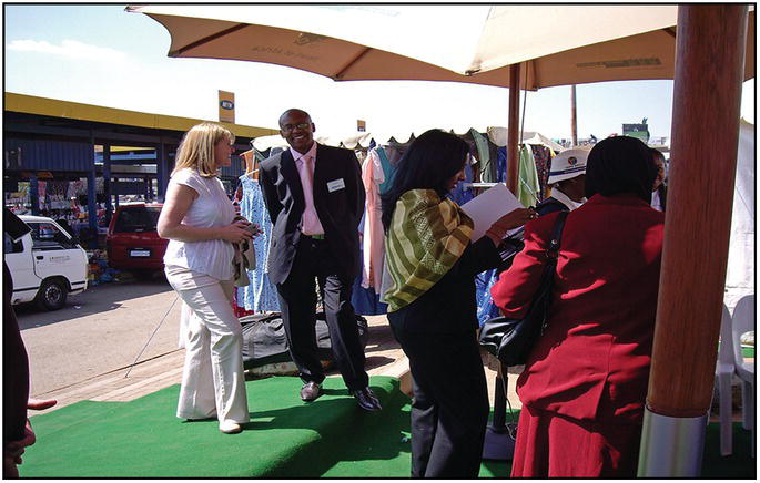 Photo of an African man in suit smiling at the camera while a Caucasian woman talks to him in a bazaar.