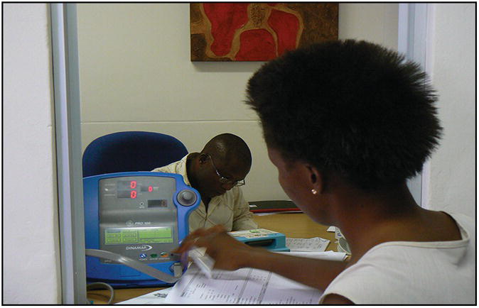 Photo of an African woman operating a DINAMAP equipment while a man in the background works on a document.