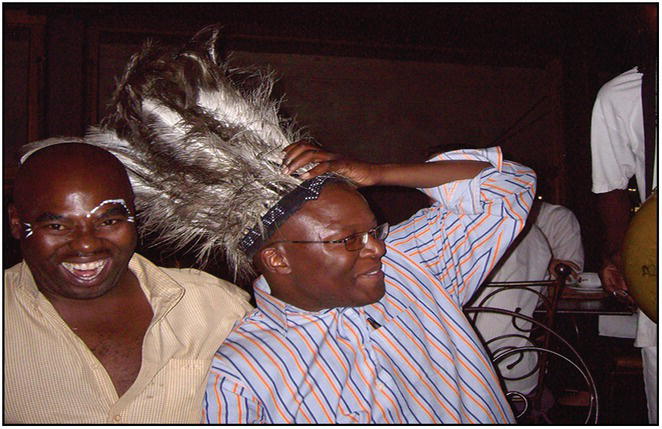 Photo of two African men. The one with a face paint smiles to the camera while the other holds onto his headdress.