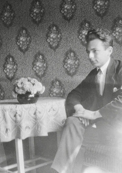 Foto Kästners aus der Schulzeit. Er sitzt an einem Tisch mit gemusterter Tischdecke und einer Blumenvase vor einer großformatig geblümten tapezierten Wand.