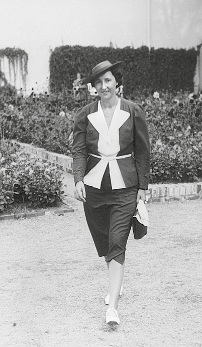 Foto von Ilse Julius im dunklen Kostüm mit heller Bluse, einem flachen Hut und einer Handtasche beim Spaziergang. Im Hintergrund sind Blumenrabatten zu sehen.