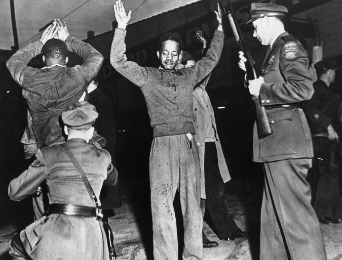 African American men hold up their hands as police with guns pat the men down.