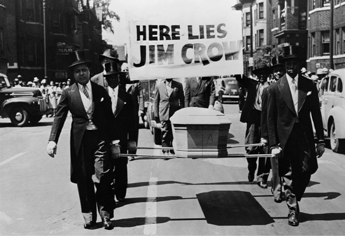 African American men in tuxedos walk down the street carrying a coffin and a sign that reads: “Here Lies Jim Crow”