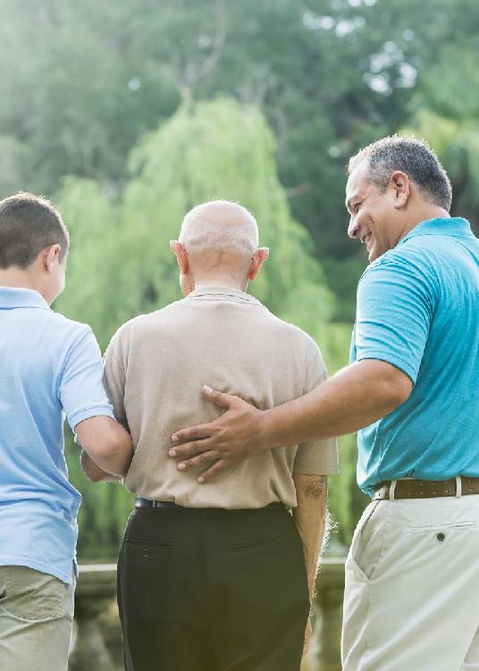 A photo showing a father and son holding onto an elderly man looking over a small bridge at the scenery across the way.