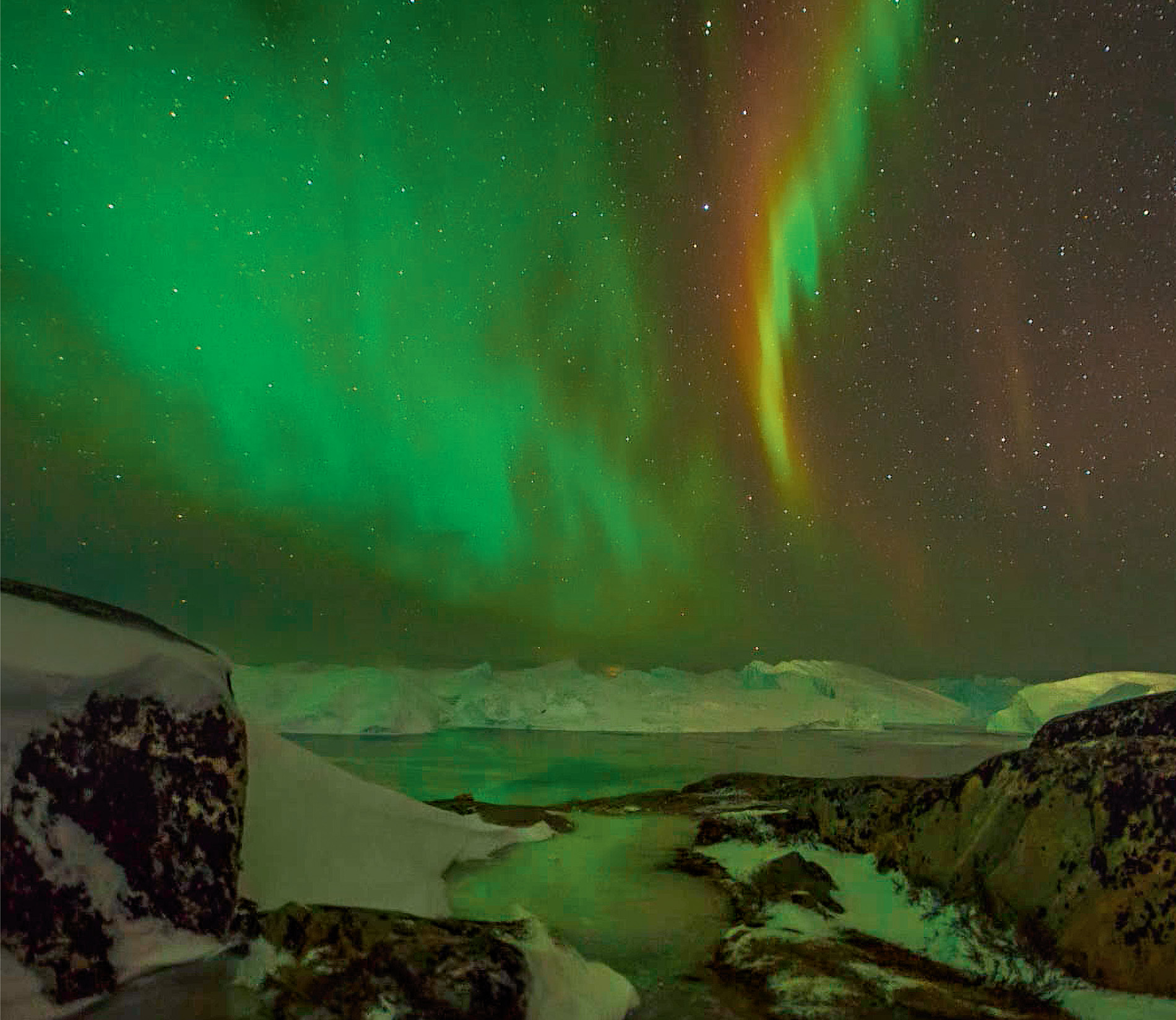 The waving green and red curtain of the Aurora Borealis is reflected in the ice and snow on the Greenland coast.