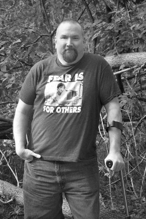 A picture of the author, E.H. Vick, in the woods. He is wearing jeans, a Bruce Lee "Fear is for others" T-shirt and using a Canadian crutch for support.