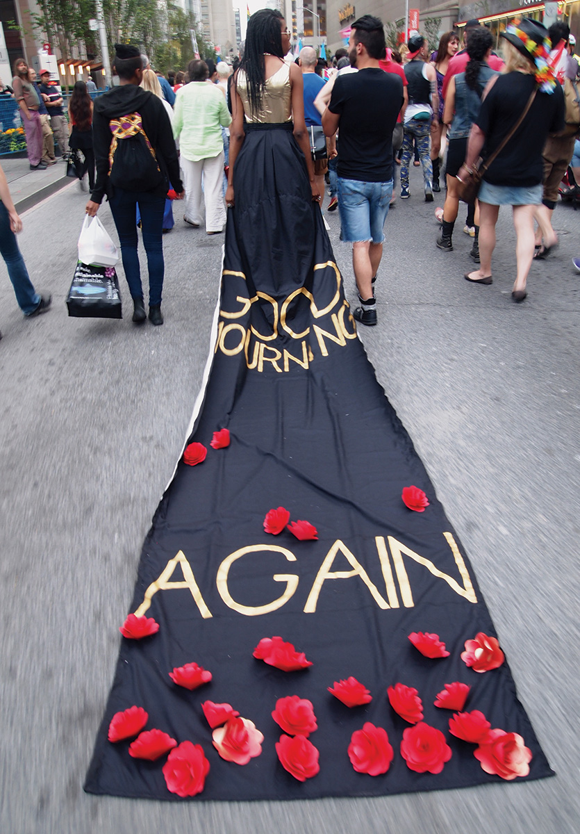 In the evening light, Luna Saint Laurent is walking away from the camera, amidst a large group of people, in the street. A long train flows back from her dress, from below a bodice, towards the camera behind her. The train is black and drags on the ground. Near where the train begins to touch the ground behind Luna’s feet is written in bold lettering “GOOD MOURNING” and then near the end of the train in the foreground, is written “AGAIN,” surrounded by paper roses.