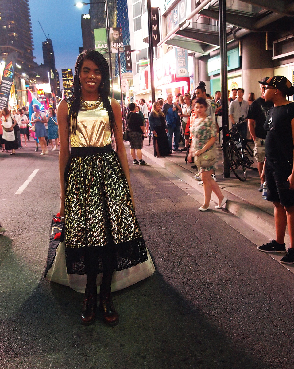 It is dusk, and Luna Saint Laurent, a Black woman with long braids and a bright smile, is standing in the foreground, wearing a dress made of reflective gold on top and the same on the bottom but covered by a layer of black geometric lace. On her feet are combat boots. A long train follows behind her. She is standing on the street, and behind her to her right is a small crowd walking forward, holding flags and large signs. There is also a crowd of people standing on the sidewalk behind her to her left walking and watching the march. Buildings, signs, bright lights, and a cityscape frame the top of the image.