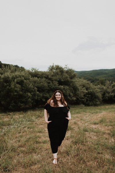 Lara is dressed in a black romper with her hands tucked into her pockets. Her brown curly hair is down in front of her shoulders. She’s standing in a field with the woods behind her. She is smiling in this photo.