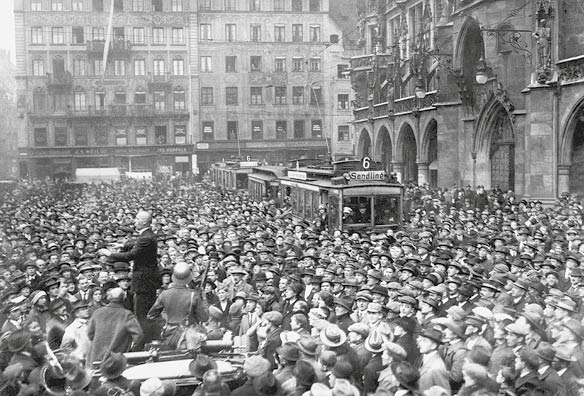 Menschenmasse am M¸nchner Marienplatz. Rechts vor dem Rathaus zwei Trambahnen der Linie 6 Richtung Sendling, die wegen der Menge der Schaulustigen anhalten mussten. Links auf einer Kutsche steht Julius Streicher und h‰lt eine Rede.