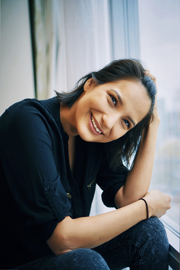 A young Indigenous woman smiling at the camera. She is sitting with her elbows propped on her knees and is resting her head on her left hand.