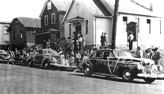 A photo of a group of people standing outside the one story Baptist Church. Cars are parked outside the church.