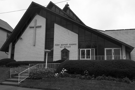 A photo of the one story building of the First Baptist Church of Nutley. The building has sloping roof, and a cross is mounted the wall facing the street.