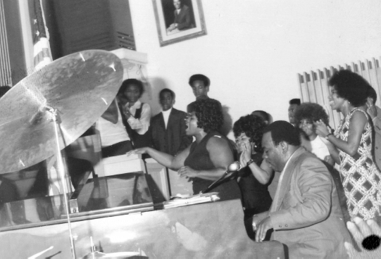 A photo of Reverend Lawrence Roberts playing the piano and Gertrude Hicks singing. Several people in the room clap to their performance.