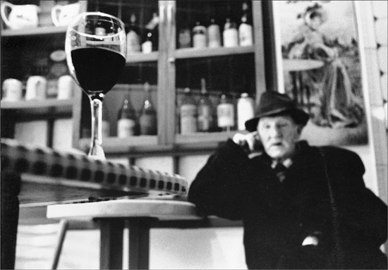 An older gentleman staring down a bar at a glass of red wine