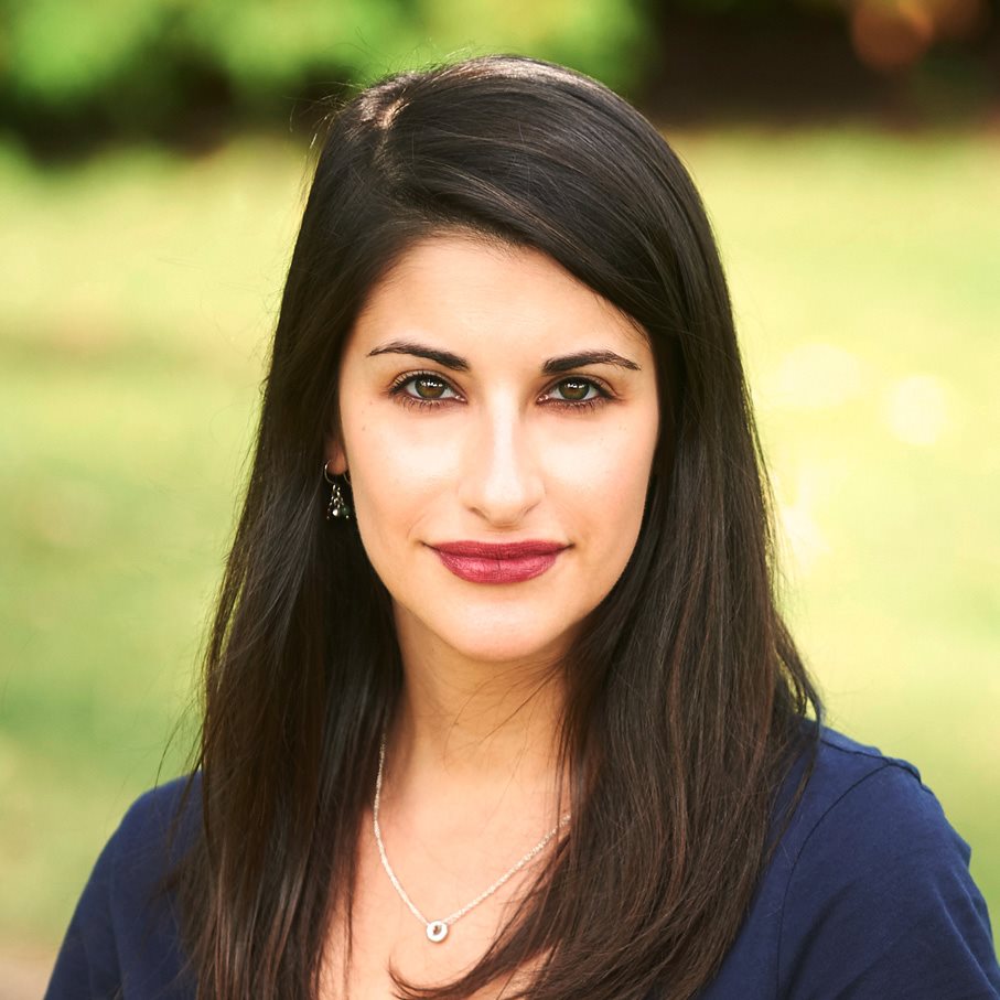 Jordi looks directly at the camera, framed from her head to her shoulders in a square border with an unfocused green background. She is in her thirties, has long black hair and an oval face. She smiles slightly at the camera.