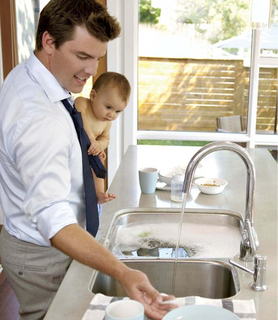 Photo of a father holding his baby in one hand while he washes dishes with the other.