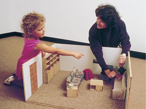 These three photos show the following: 1. An adult hides a toy in a small-scale, dollhouse-sized room. 2. The child finds a similar toy in the full-sized room, using the small-scale room as a model for where to find it. 3. The child also finds the toy in the same place in another small-scale model room.