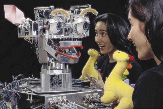 Photo of a designer of the robot Kismet interacting with it as though it were a baby. The robot and human are making eye contact, and the human is holding a stuffed dinosaur.