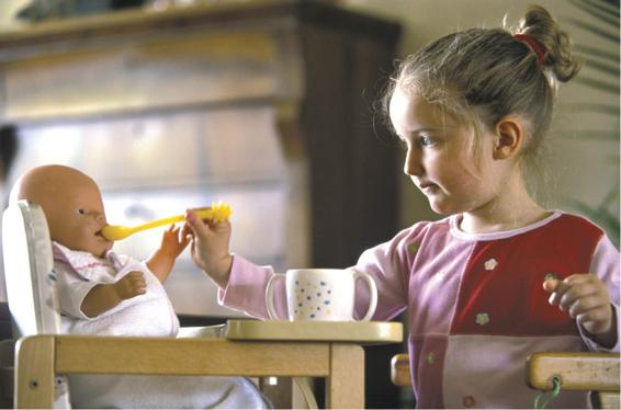 Photo of girl pretending to feed a baby doll that is seated in a high chair.