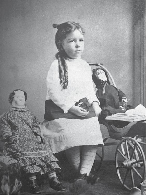 Photo of a little girl, probably around 1900 to 1910, sitting quietly with a doll in a chair on one side, and a doll in a carriage on the other.