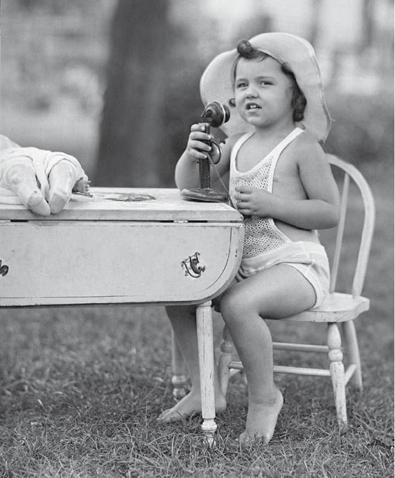 Photo of a little girl, probably around 1950, sitting at a table with a doll on it, pretending to talk into a telephone or microphone.