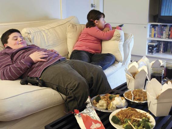 Photo of two chubby children sitting on the couch. One is watching television and changing channels. The other is leaning back and does not appear to be engaged in any activity. The coffee table in front of them is filled with take-out food.