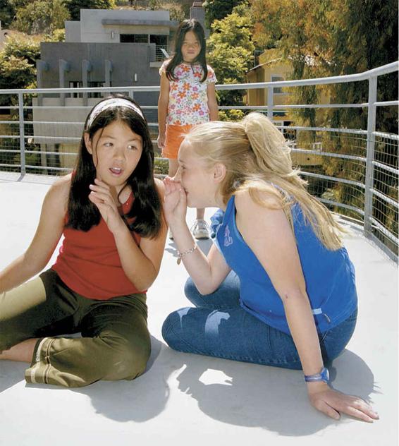 Photo of two girls whispering and giggling together while a third girl is excluded and looking on, probably the subject of the girls’ whispering.