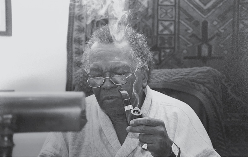 Photograph shows Austin Clarke writing. He's seen smoking a pipe.