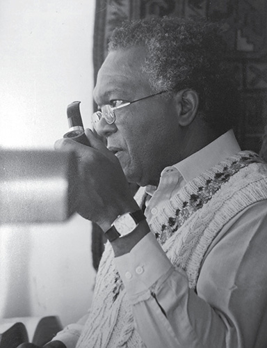 Photograph of Austin Clarke looking out a window, as he holds a smoking pipe.