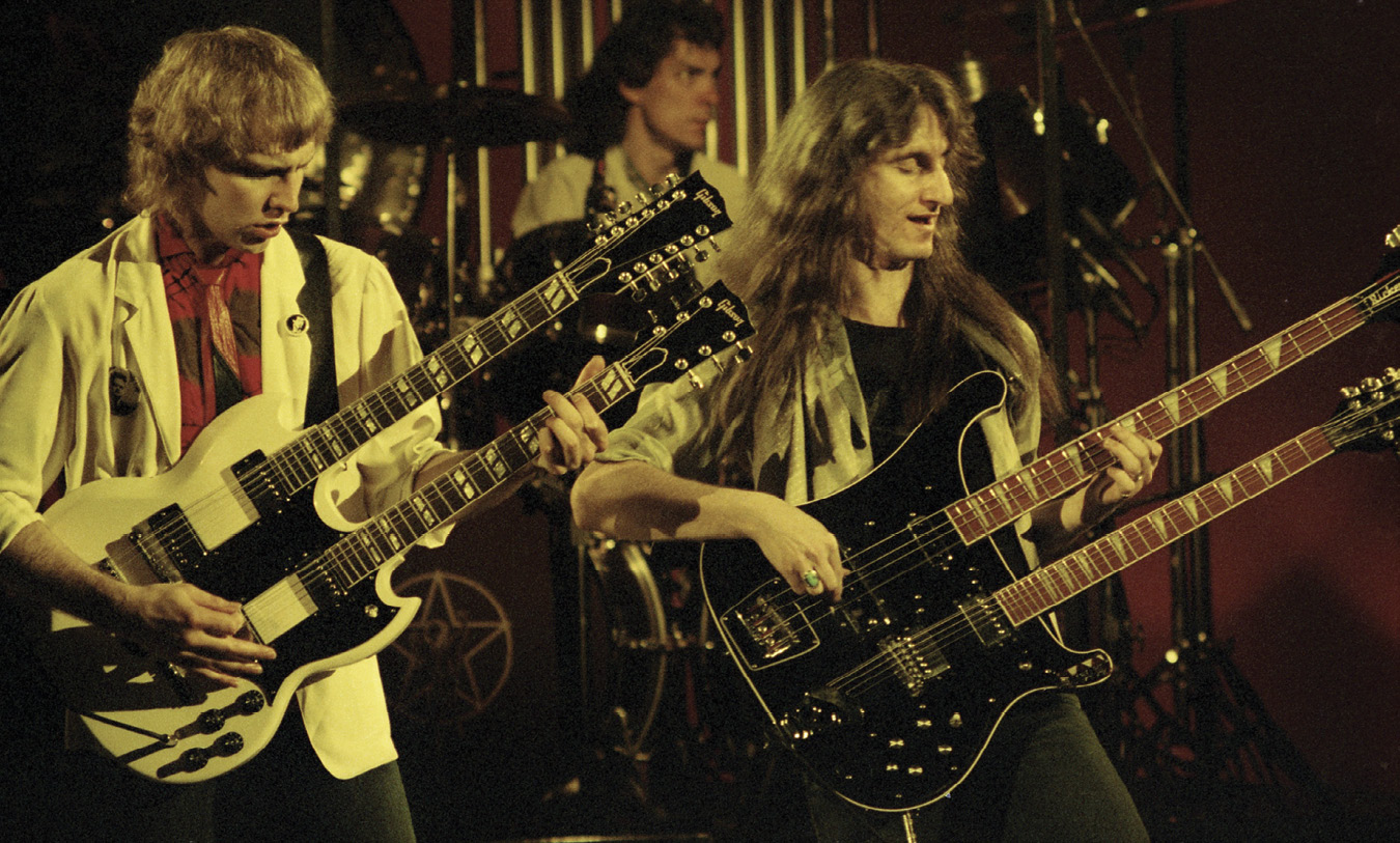 Alex, playing a white double-necked guitar, and Geddy, playing a black double-necked bass, with Neil in the background.