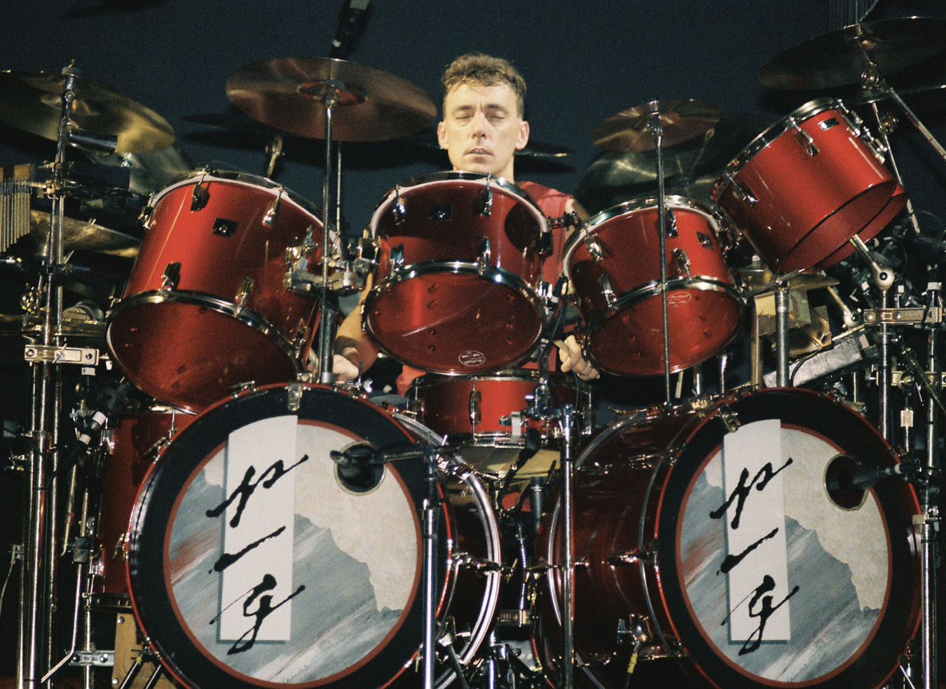 Neil, with a crew cut, seated behind a red drum kit. Text on the bass drums read 'P/G.'