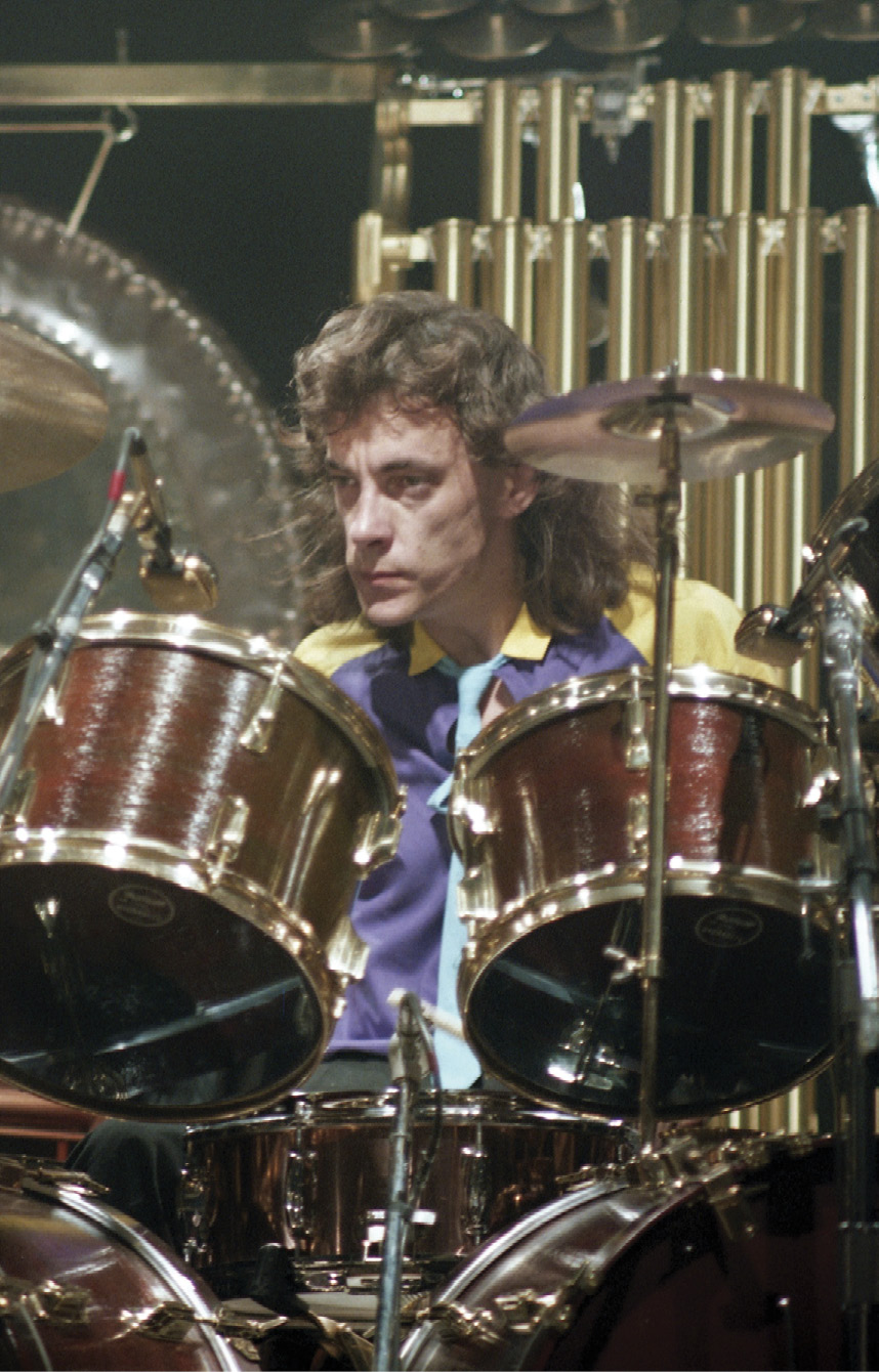 Neil, with long hair, sits behind a drum kit. He's wearing a yellow and purple shirt with a light blue tie.