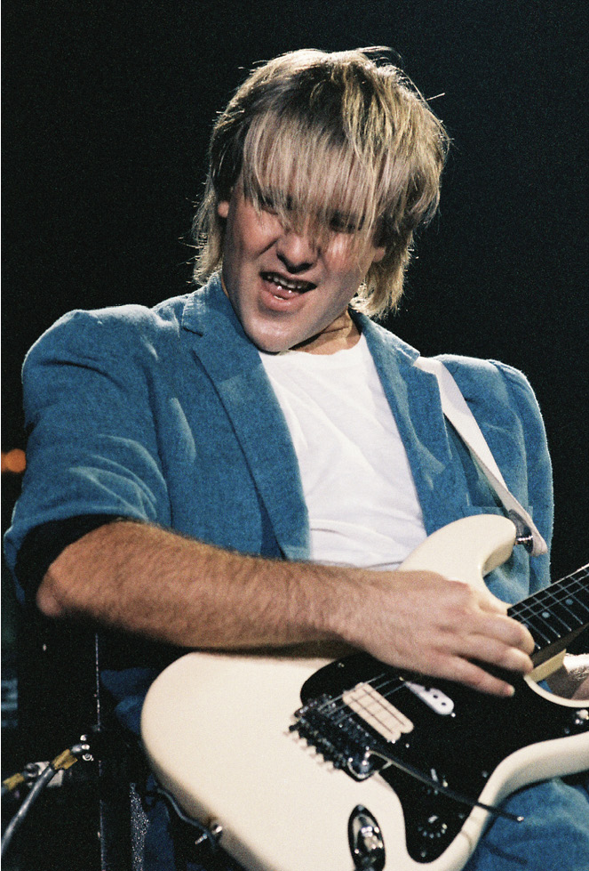 Alex, wearing a blue suit, playing a white guitar.