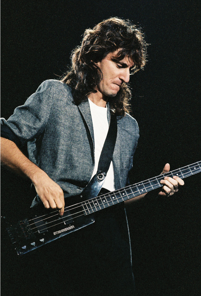 Geddy, wearing a grey suit jacket, playing the bass guitar.