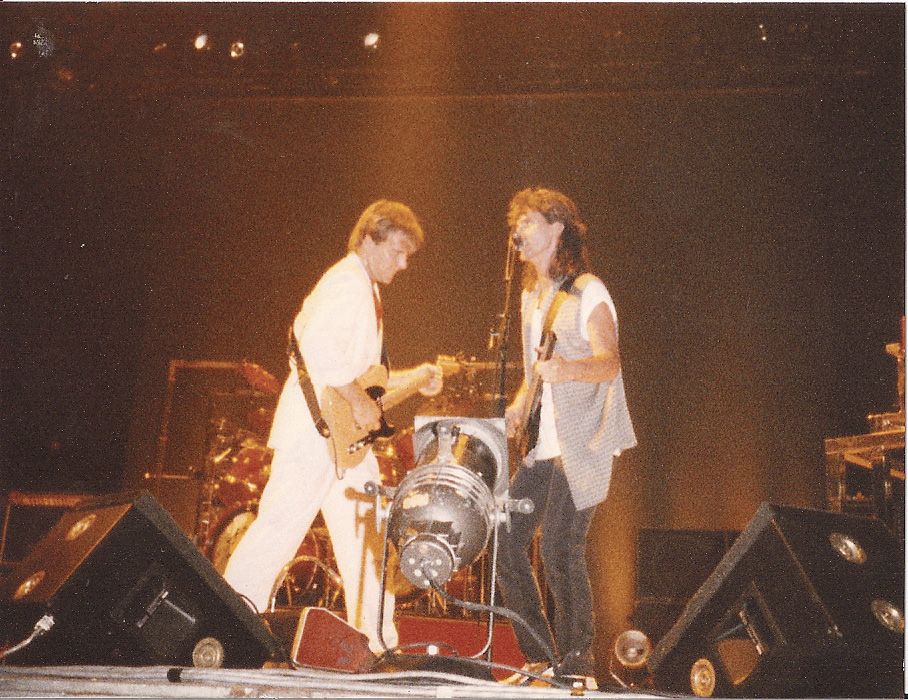 Alex and Geddy perform on stage next to each other. Alex, on the left, plays his guitar, while Geddy, on the right, sings into the microphone as he plays his bass guitar.