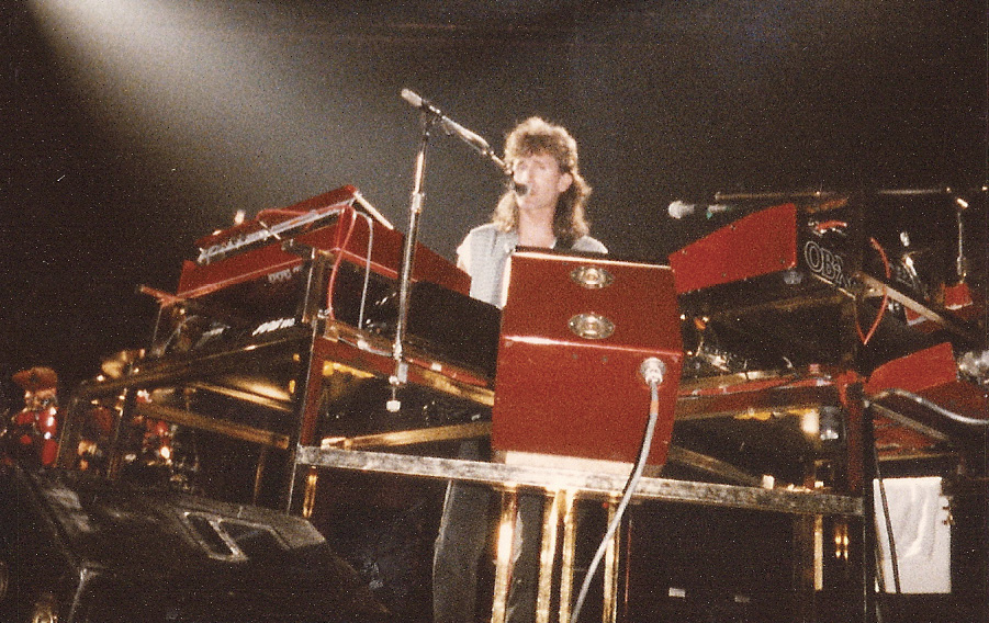 Geddy sings into a microphone as he plays the keyboard.