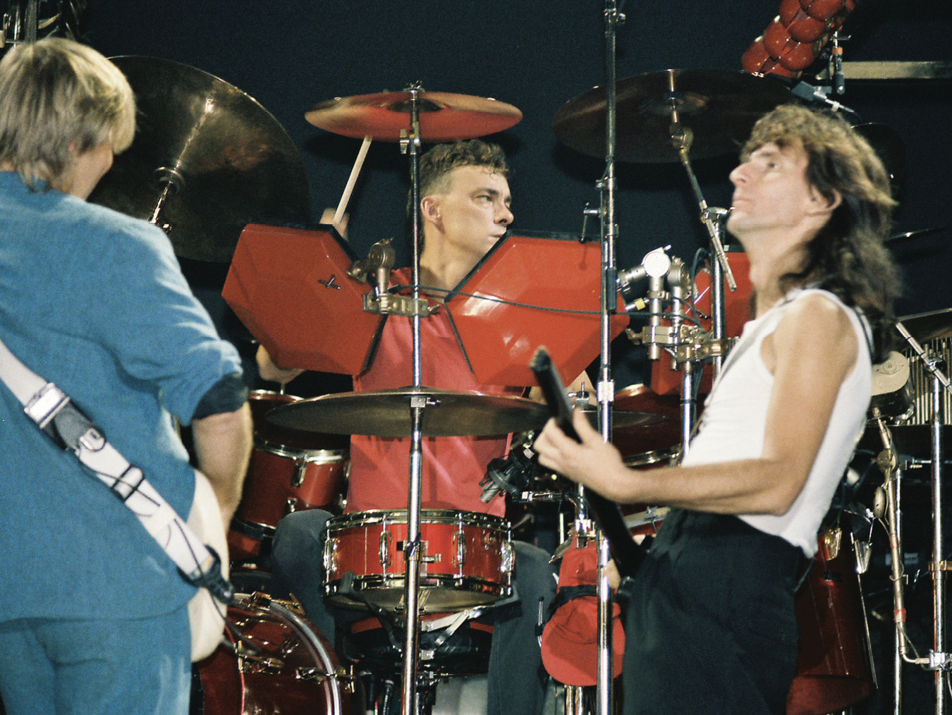 Alex, Geddy, and Neil on stage with their instruments. Alex has his back to the camera and faces Neil, who's seated behind a red electronic drum kit. To the right of them, Geddy is playing his bass guitar as he looks off into the distance beyond the camera's view.