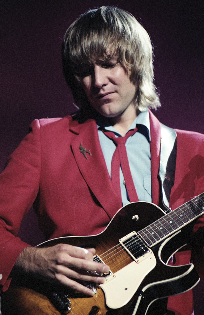 Alex, wearing a red suit jacket and a light dress shirt with a medium-length 'new wave' haircut, plays an electric guitar.