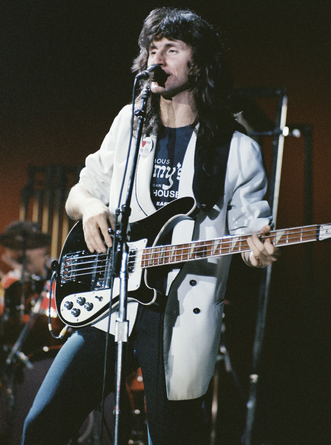 Geddy, wearing a long white coat, sings into a microphone while playing an electric bass guitar.