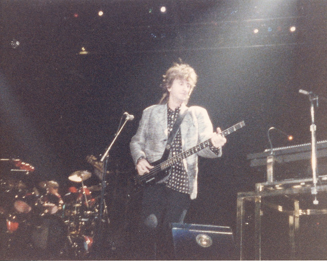 Geddy, in a grey suit jacket, playing the bass guitar.