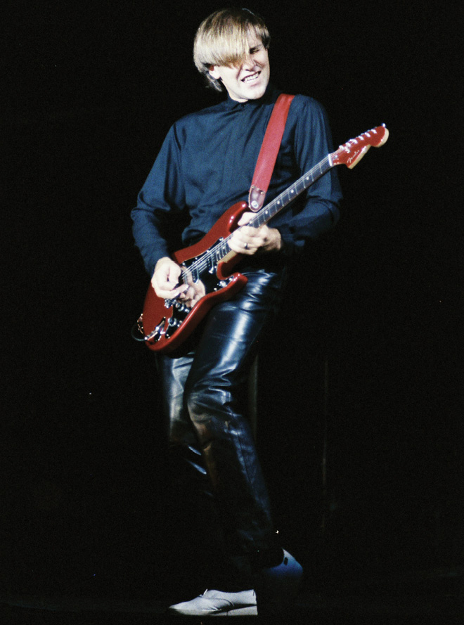 Alex, wearing a black shirt and black leather pants with a 'pageboy' haircut, plays a red electric guitar. He grimaces as he strums his guitar.