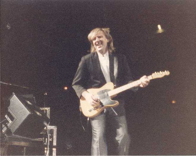 Alex, wearing jeans and a suit jacket, playing the guitar.