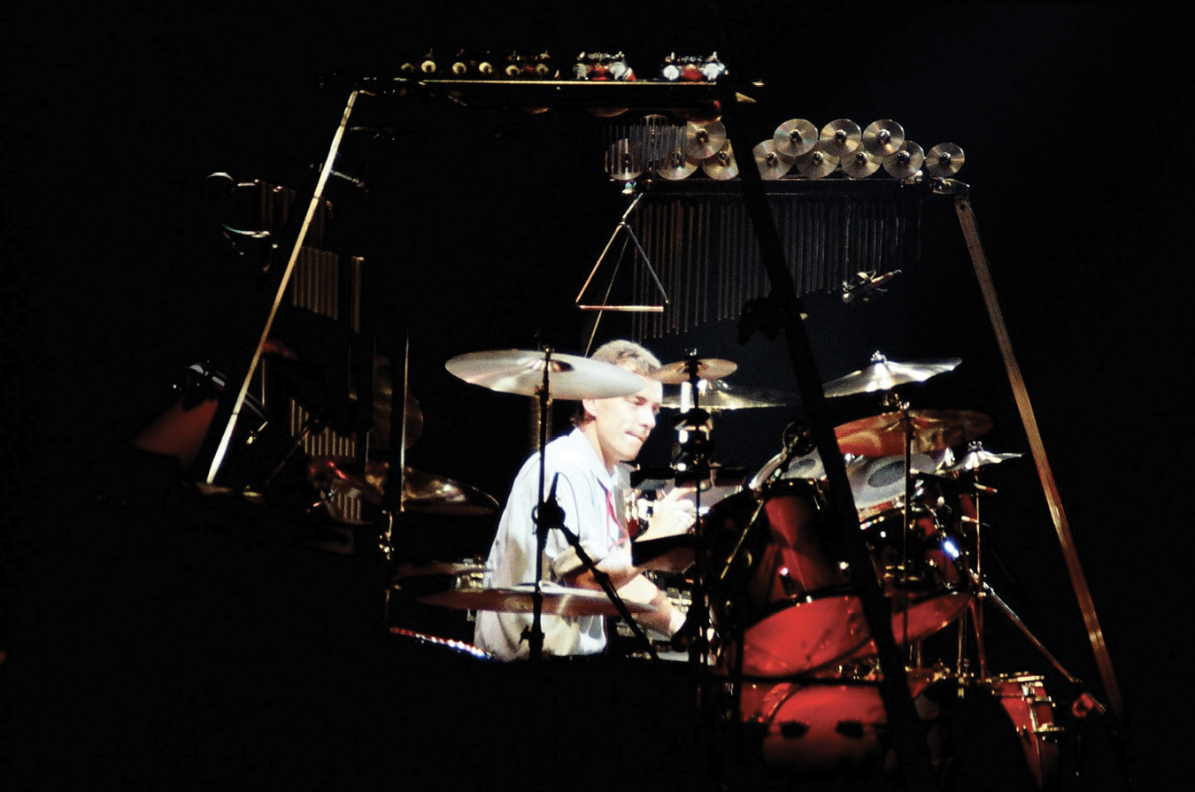 Neil, with short hair, playing percussion. He is surrounded by a drum set and other percussive instruments, including bells, a triangle, and an array of smaller cymbals to his side.