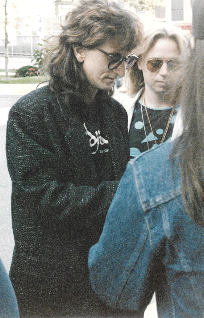 Geddy, wearing sunglasses and a black jacket, signs autographs for two fans outside.