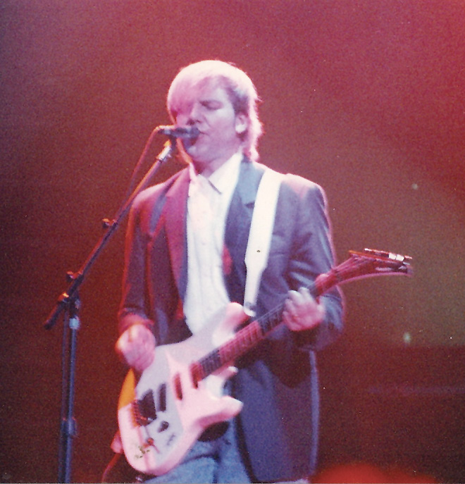 Alex, wearing a blue suit jacket and a white shirt, sings into a microphone as he plays his guitar.