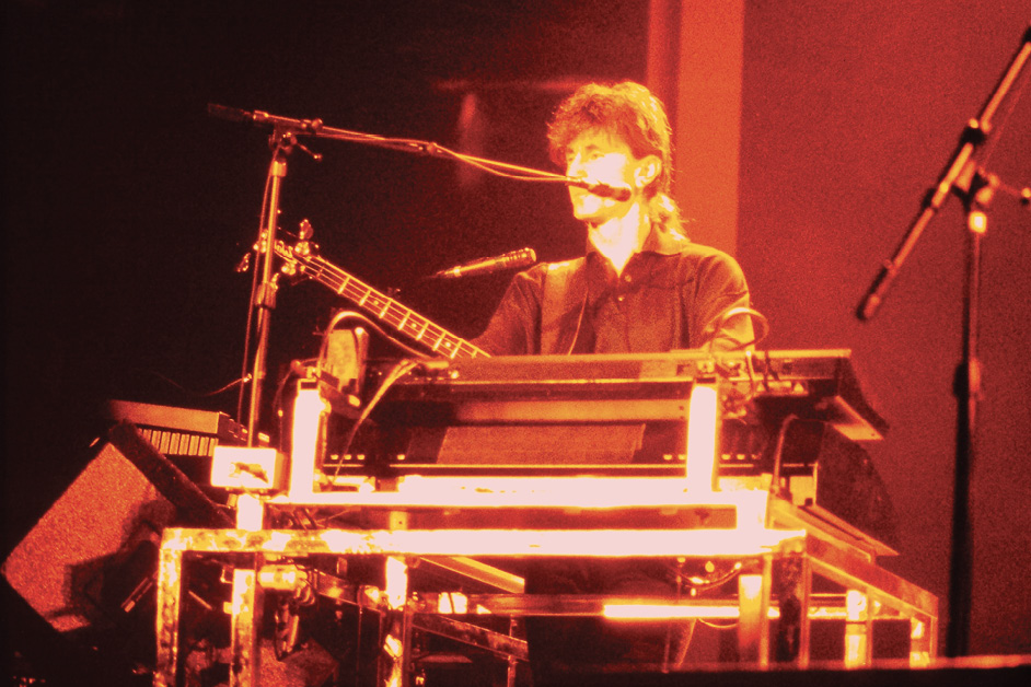 Geddy, with his hair in a pony tail, plays the keyboard on stage as he's illuminated by the stage lights.