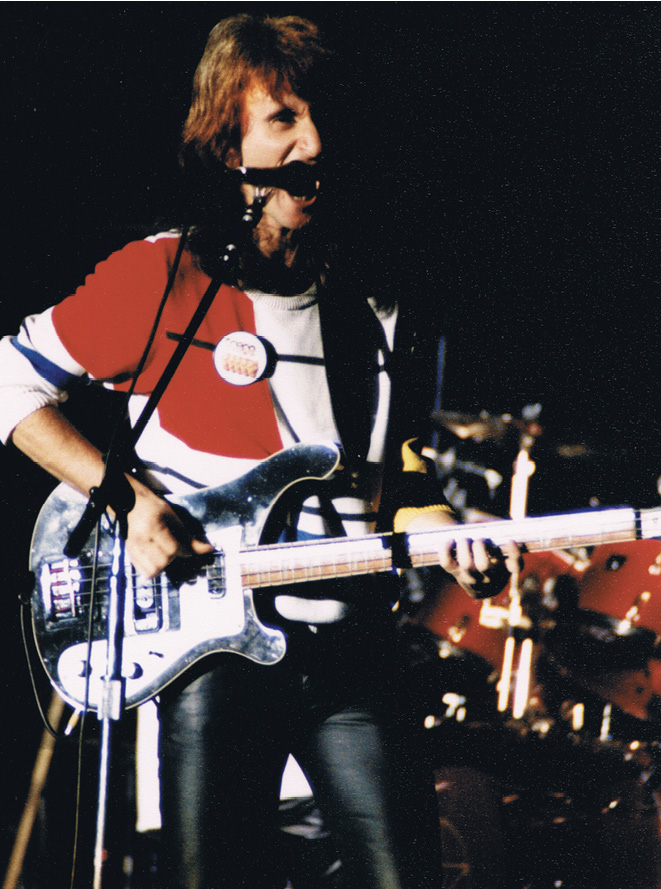 Geddy, wearing a red and white shirt with a large button, playing a black bass guitar.