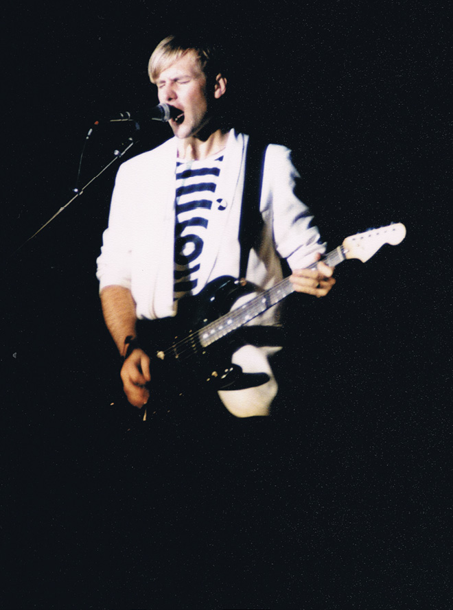 Alex, wearing a white sports coat with a black and white t-shirt, sings into a microphone.